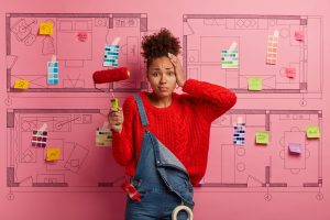 Puzzled nervous dark skinned woman has displeased face expression, holds paint brush, tired of house refurbishment, stands against house sketch, thinks about room improvement. Repair, housework